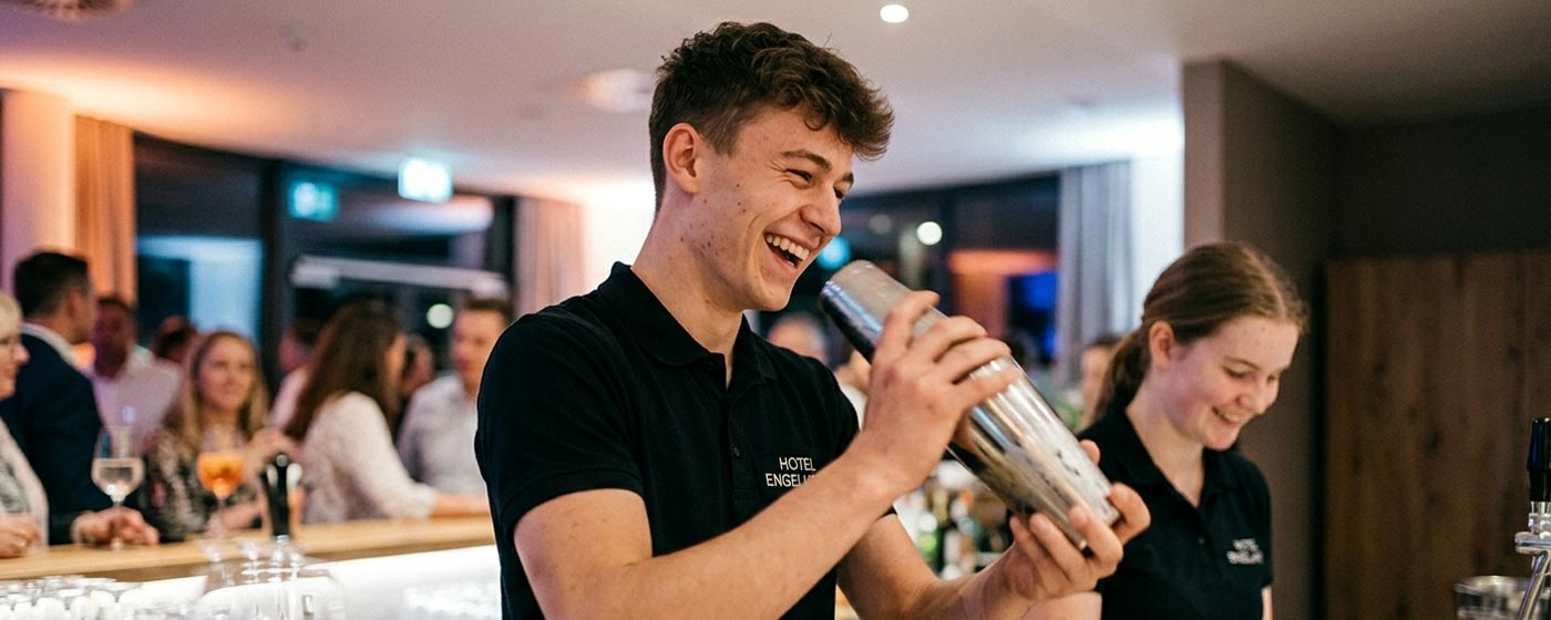Lachender junger Mitarbeiter im Engelhorn-Polo mixt einen Cocktail im Shaker, rechts eine Kollegin, dahinter Gäste an der Bar