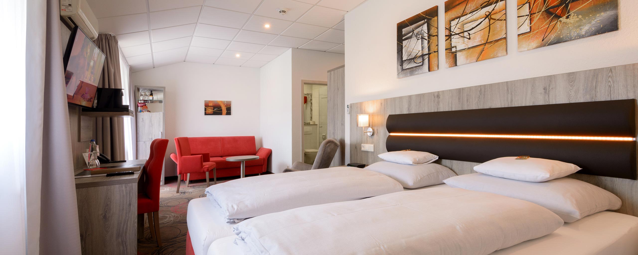 Hotelzimmer mit zwei Betten vor dunklem Kopfteil mit Lichtleiste, roter Sitzgruppe am Fenster, Schreibtisch und Wandfernseher