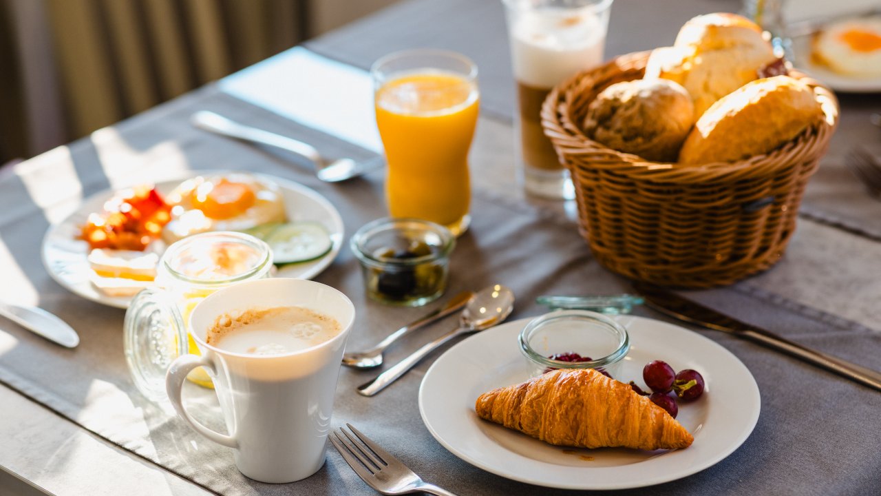 Gedeckter Frühstückstisch im Sonnenlicht: Croissant mit Trauben, Latte Macchiato, Orangensaft und ein Korb mit Brötchen