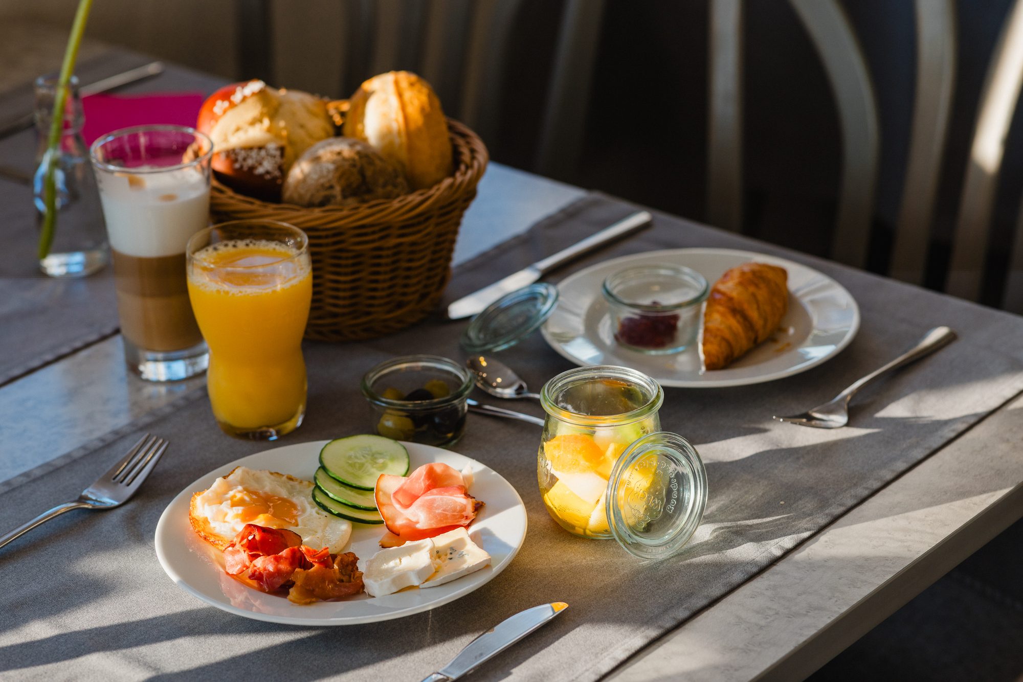 Gedeckter Frühstückstisch mit Brötchenkorb, Latte Macchiato und Orangensaft im Sonnenlicht